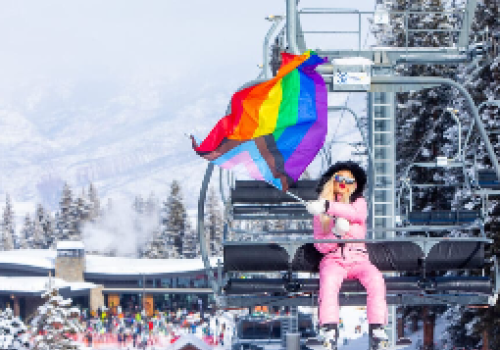 A person in pink ski gear is riding a chairlift, waving a rainbow flag amidst snowy mountains and skiers below.