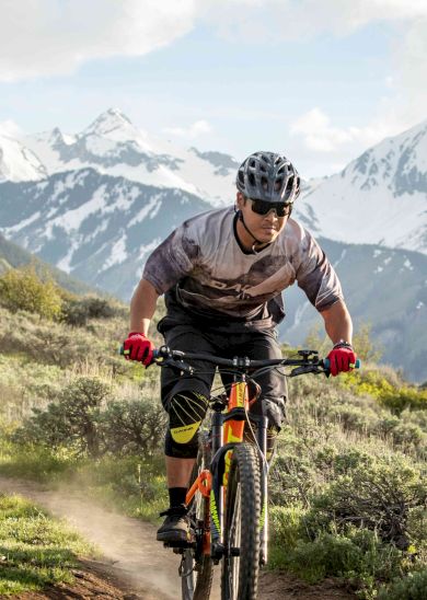 A person mountain biking on a trail surrounded by lush greenery and snow-capped peaks under a partly cloudy sky, wearing a helmet and sunglasses.