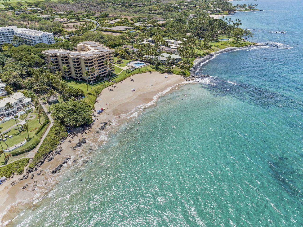 Aerial view of a coastal area with a sandy beach, turquoise water, rocky shoreline, and nearby buildings surrounded by lush greenery.
