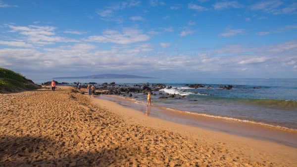 A sandy beach with people walking along the shoreline, blue sky, and scattered clouds. There are rocks in the water and distant land visible.