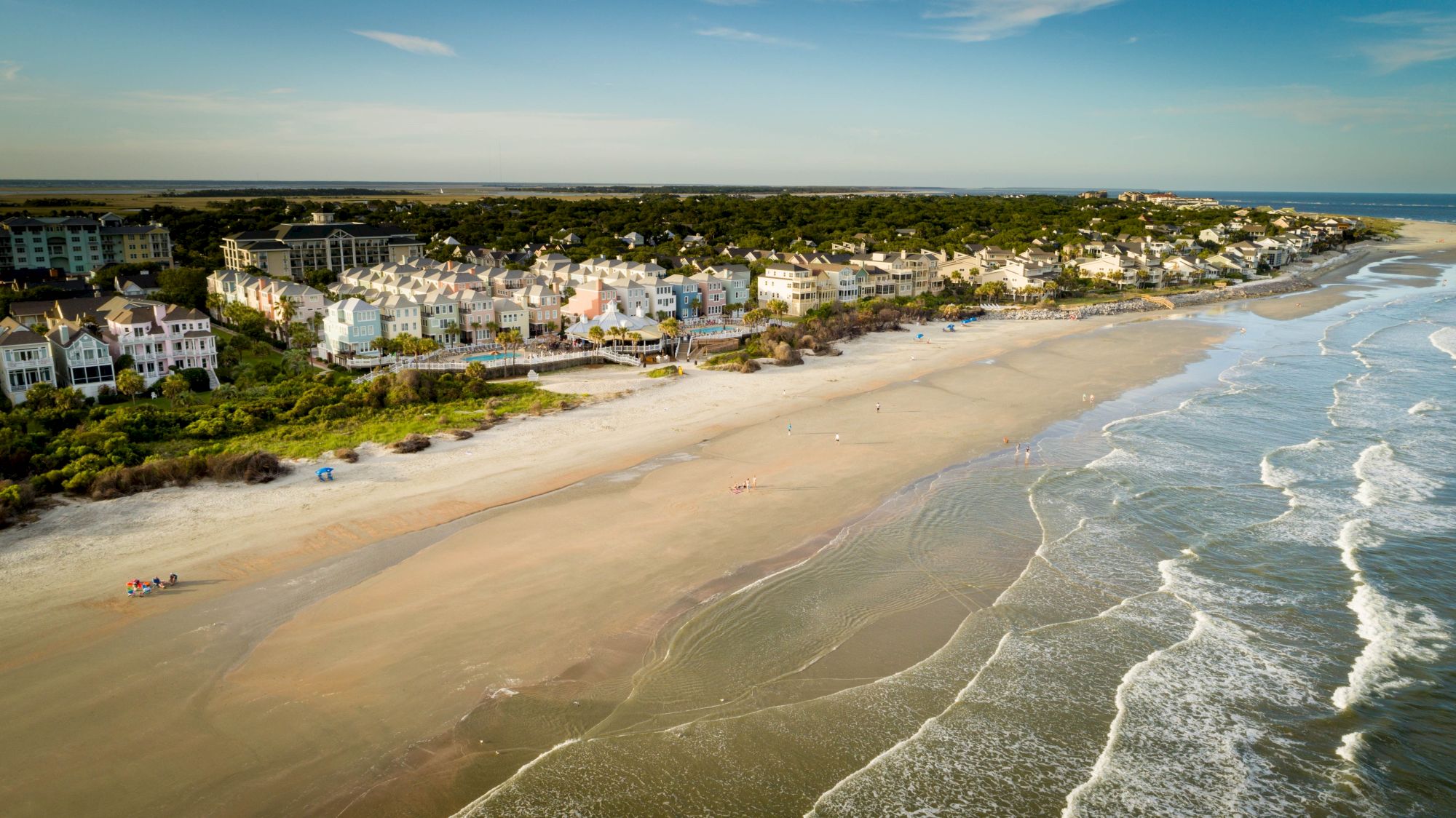 An aerial view of a coastal town with houses lined along the beach, waves gently hitting the sandy shore, and a clear blue sky above.