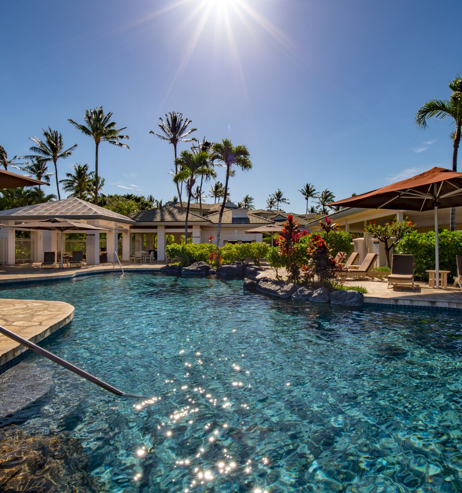 A tropical pool scene with clear blue water, umbrellas, and palm trees under a bright sun.