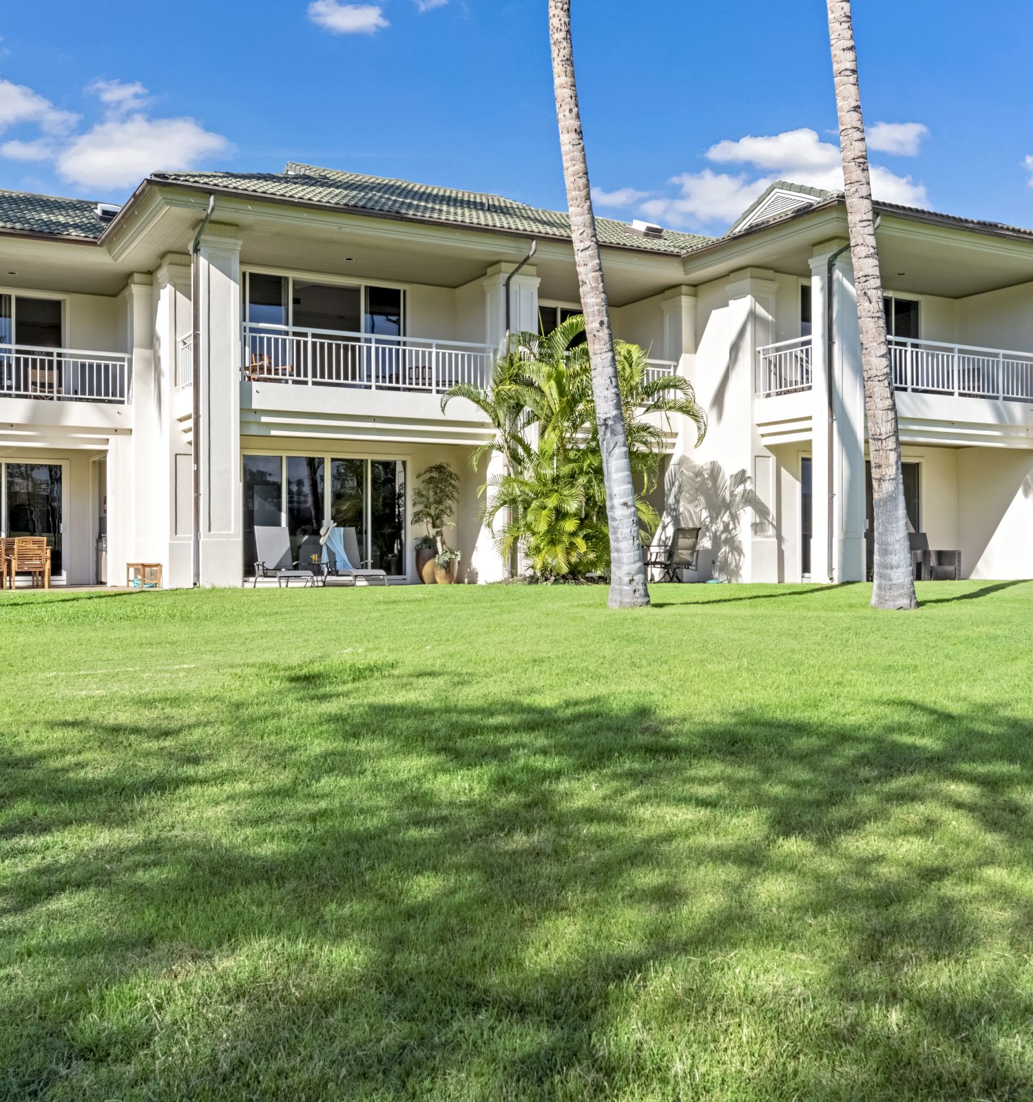 The image shows a large house with balconies, surrounded by a grassy lawn and palm trees under a clear blue sky.