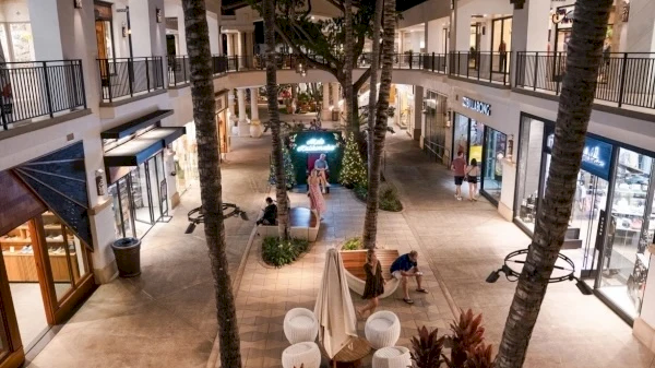 An outdoor shopping mall with palm trees, stores, and people walking and sitting around. It appears to be evening with lights on.