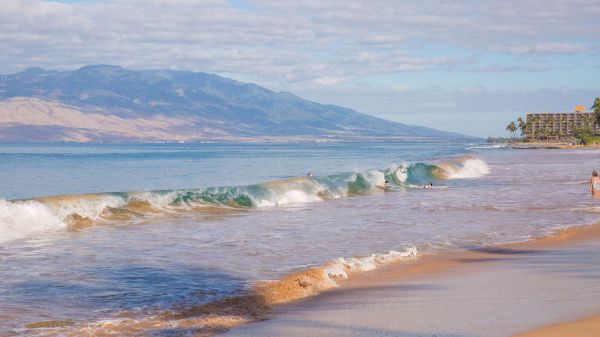 A sandy beach with gentle waves, distant mountains, and buildings on the right under a partly cloudy sky.