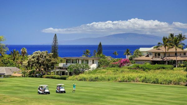 A golfer on a lush course, with golf carts. Scenic view includes houses, ocean, and mountains under a bright blue sky with clouds.