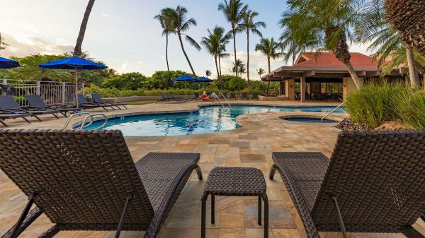 Two lounge chairs face a serene pool surrounded by palm trees and a patio with blue umbrella shades.