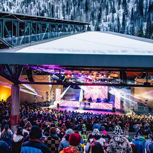 A lively outdoor concert with a large crowd, vibrant stage lights, and a snowy mountain backdrop, under a modern pavilion.