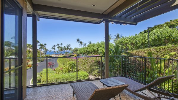 A balcony with lounge chairs overlooks lush greenery, a road, and a distant ocean view under a clear blue sky.
