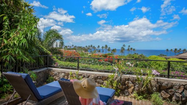 The image shows a scenic outdoor patio with lounge chairs, a person wearing a hat, lush greenery, and a view of the ocean under a blue sky.