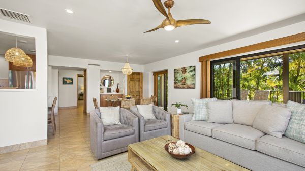 A modern living room with gray sofas, a wooden coffee table, and ceiling fan, featuring large windows overlooking greenery outdoors.