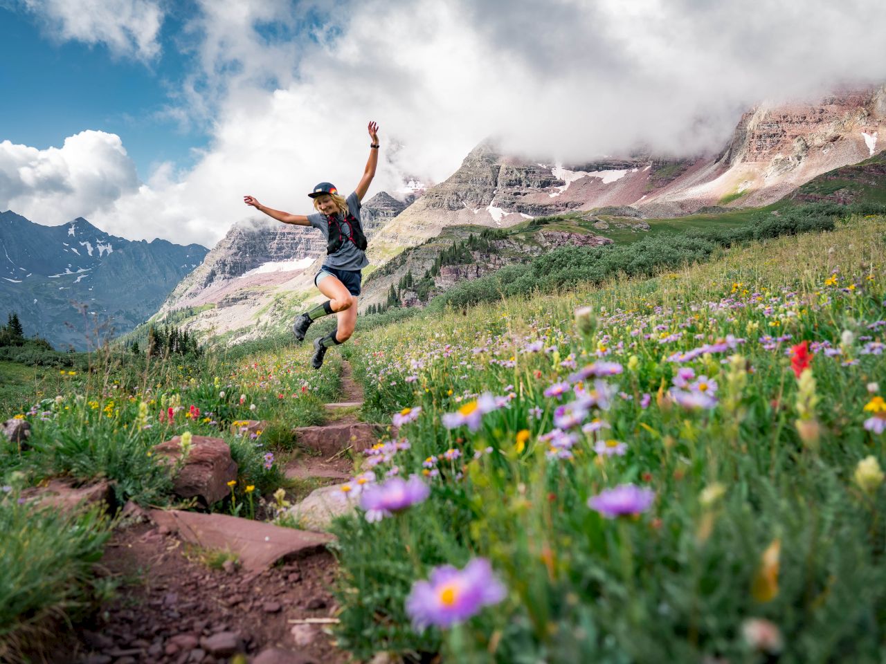 A person joyfully jumps on a mountain path surrounded by vibrant wildflowers, with misty mountains in the background under a cloudy sky.