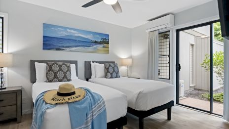 The image shows a bedroom with two single beds, a hat, and a shawl on one bed. There’s a ceiling fan and a sliding door leading outside.