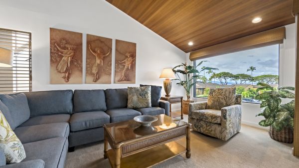 A cozy living room with a grey sectional, patterned armchair, wooden ceiling, and large window showcasing a scenic outdoor view.
