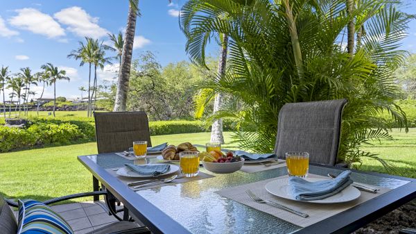 An outdoor dining table with plates, napkins, glasses of orange juice, fruit, and croissants. Tropical greenery and palm trees in the background.