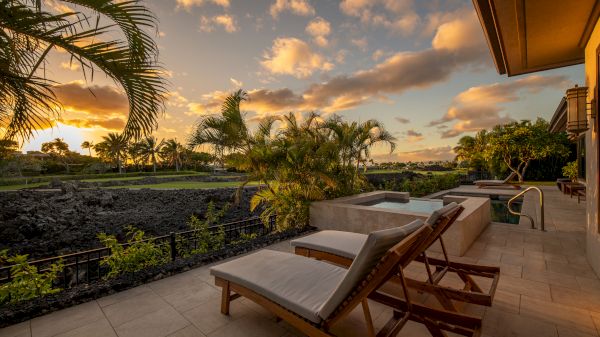 A tranquil patio features lounge chairs, a pool, lush greenery, and a scenic view of the sunset with palm trees and a volcanic rock landscape in the distance.