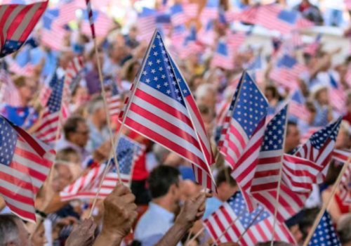 A crowd of people waving American flags, creating a festive and patriotic atmosphere at what seems like a public gathering or event.