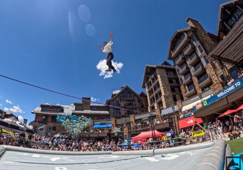 A person is performing a high jump onto an inflatable airbag in a busy outdoor setting with buildings and a crowd below.