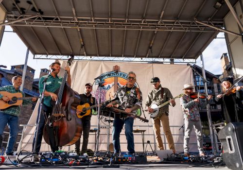 A group of musicians performing on an outdoor stage with various instruments, and a banner backdrop displaying colorful graphics and text.