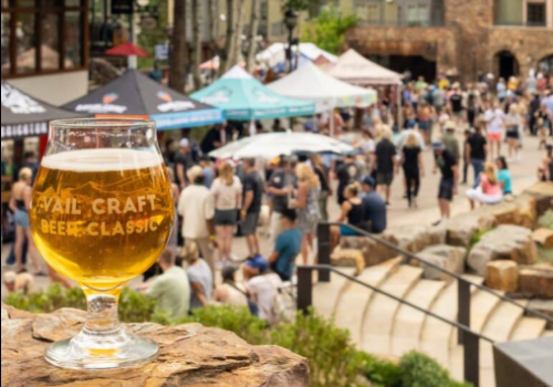 A glass of beer sits on a rock with a crowd and tents in the background at an outdoor event.