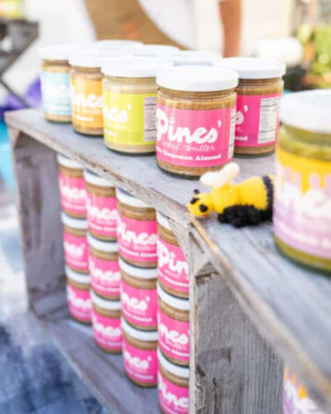 Jars of nut butter are displayed on wooden shelves, alongside a small toy bee decoration, creating a vibrant market stall scene.
