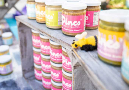 Jars of nut butter are displayed on wooden shelves, alongside a small toy bee decoration, creating a vibrant market stall scene.