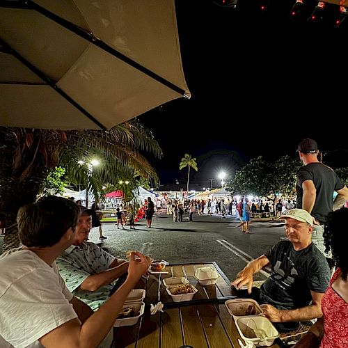 People are dining at an outdoor table under a patio umbrella at night, with a lively street market in the background.