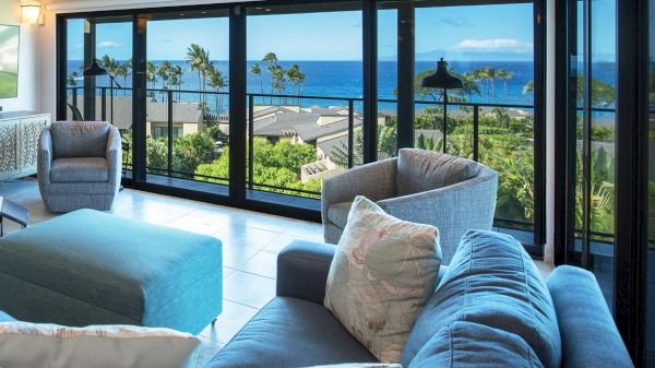 A living room with blue furniture offers a panoramic view of the ocean and greenery through large sliding glass doors.