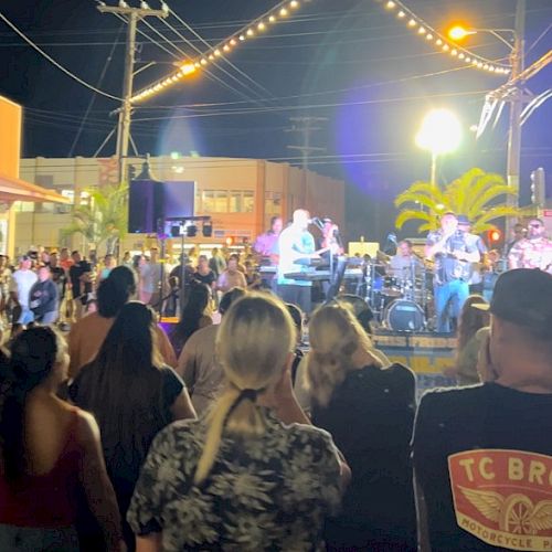 A crowd watches a live outdoor music performance at night, with string lights above, creating a lively atmosphere.