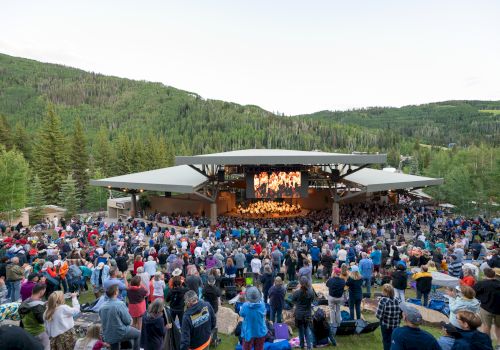 An outdoor concert with a large crowd gathered in front of a stage amidst lush, forested hills.