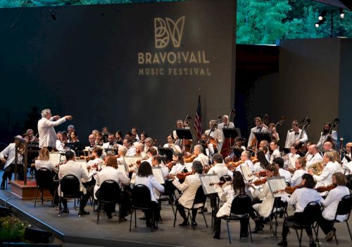 An orchestra performs on stage at the Bravo! Vail Music Festival, with a conductor leading musicians dressed in white.