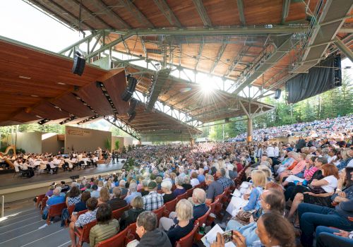 A large outdoor amphitheater with a full audience, a stage with an orchestra, and sunlight streaming through the roof structure.