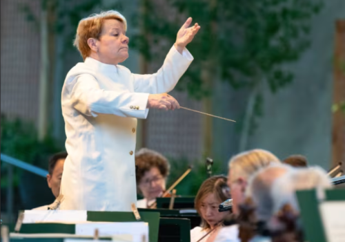A conductor in a white coat leads an outdoor orchestra performance, with musicians focused on playing their instruments.
