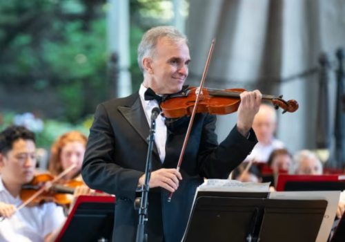 A man in a suit is playing a violin on stage with an orchestra seated behind him, in an outdoor setting.