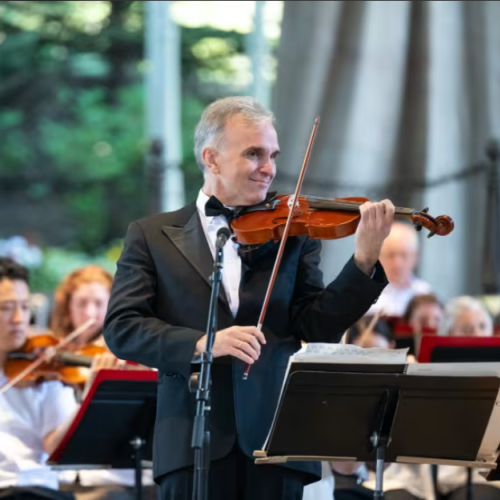 A man in a suit is playing a violin on stage with an orchestra seated behind him, in an outdoor setting.