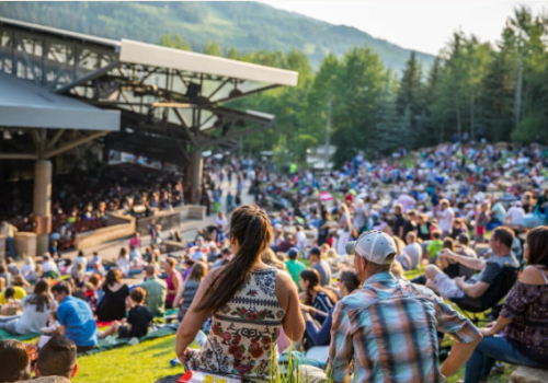 A large outdoor crowd sits on a grassy slope, enjoying a concert or performance at an amphitheater surrounded by trees and mountains.