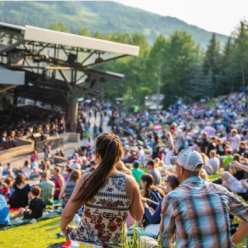 A large outdoor crowd sits on a grassy slope, enjoying a concert or performance at an amphitheater surrounded by trees and mountains.
