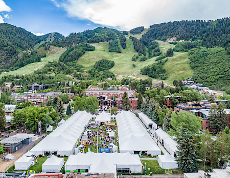 A series of white tents in a lush, mountainous area with buildings and trees, set against a backdrop of rolling green hills.