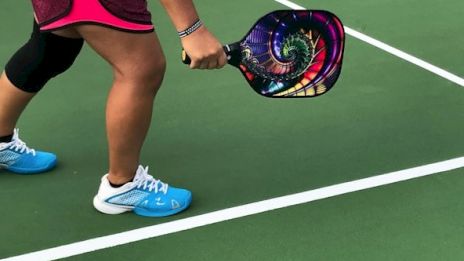 The image shows a person on a pickleball court with a colorful paddle, wearing athletic gear and shoes, ready to play.