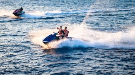 Two jet skis speeding across the water, creating splashes and waves, with clear skies and blue sea in the background.