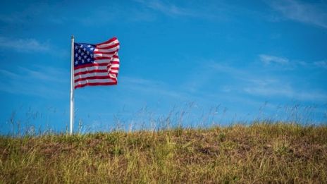 A U.S. flag on a pole flutters against a blue sky above a grassy hill, with wispy clouds enhancing the serene backdrop.
