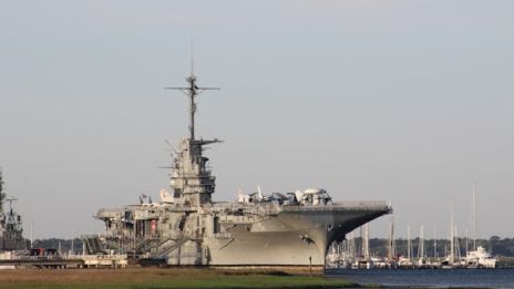 An aircraft carrier docked at a harbor with a clear sky in the background and water in the foreground.
