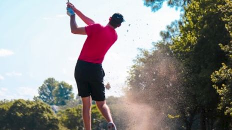A golfer in a red shirt swings a club as dirt flies from the ground on a sunny course surrounded by trees and a clear blue sky.