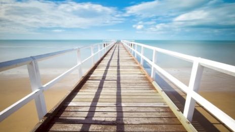 A wooden pier extends towards the horizon over a calm sea, under a partly cloudy blue sky, with shadows cast by the railings.