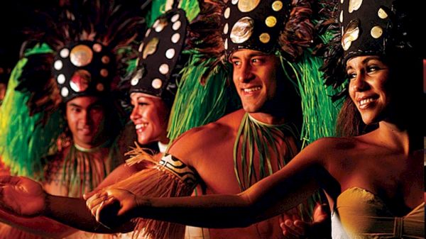 Performers in traditional attire with decorative headdresses are smiling and holding hands in a vibrant, cultural dance scene.