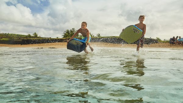 Two people running with bodyboards towards the ocean on a sandy beach, with a group sitting in the background.