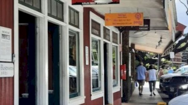 The image shows a street with shops, a red building with white windows, and people walking on the sidewalk. Signs hang above the storefronts.