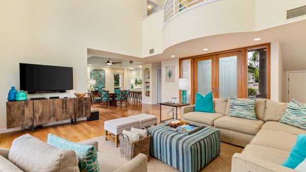 A spacious living room with beige sofas, a striped ottoman, and a TV, leading into a dining area. Ceiling is vaulted.