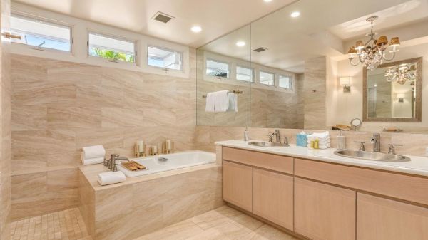 The image shows a modern bathroom with a large mirror, twin sinks, a bathtub, beige tiles, and a chandelier.