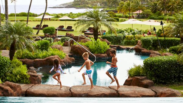 Three children run beside a lagoon-style pool in a tropical resort setting with lush greenery and ocean views in the background.